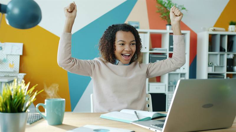 A smiling young woman raises both arms in celebration at her desk in front of a laptop.