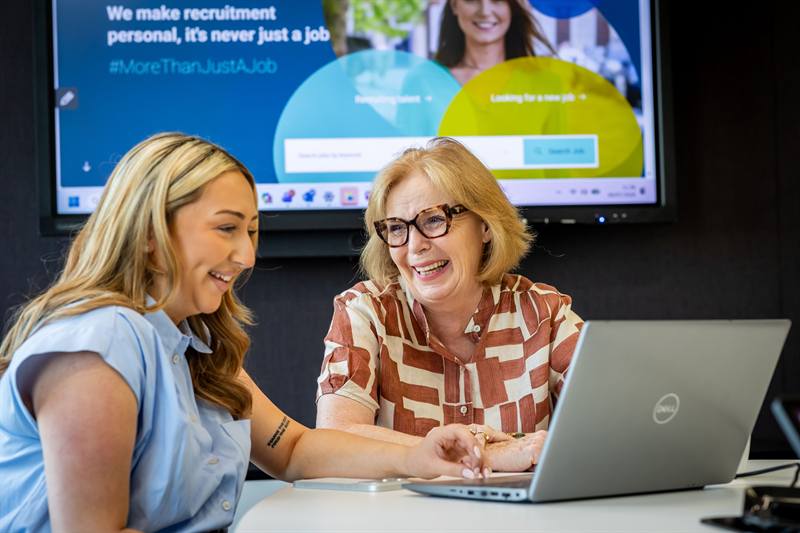 Two smiling women work together at a laptop in a meeting room.